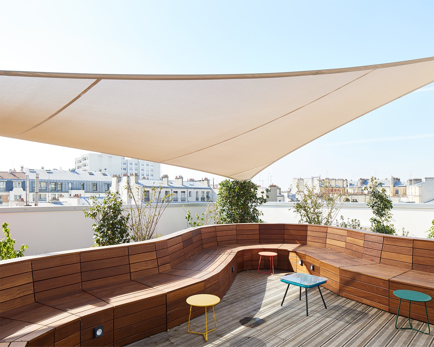 Toit terrasse en bois, mobilier coloré, banc en bois incurvé et voile blanche sur ciel bleu