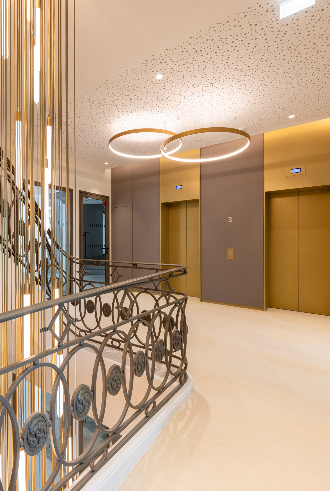 Palier du R+3 d'un immeuble de bureaux rénové avec des luminaires moderners et ascenseurs aux couleurs champagne, la rampe en ferronnerie travaillée d'un escalier.