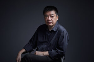 Portrait d'un homme chinois en chemise sombre sur fond sombre, légèrement de profil.